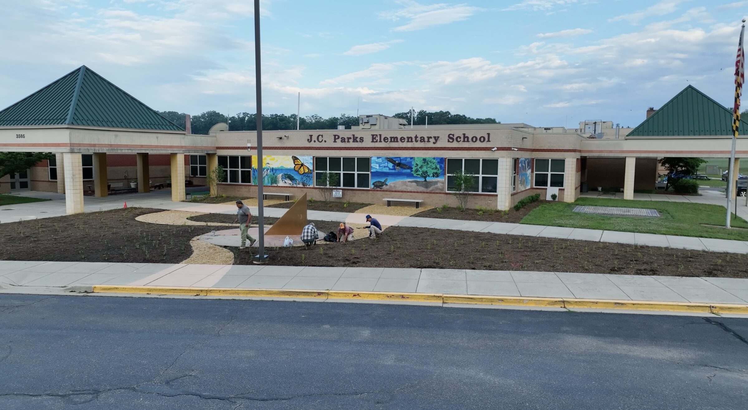 Three people working in front of J.C. Parks Elementary School, with a mural and gardens under a partly cloudy sky.